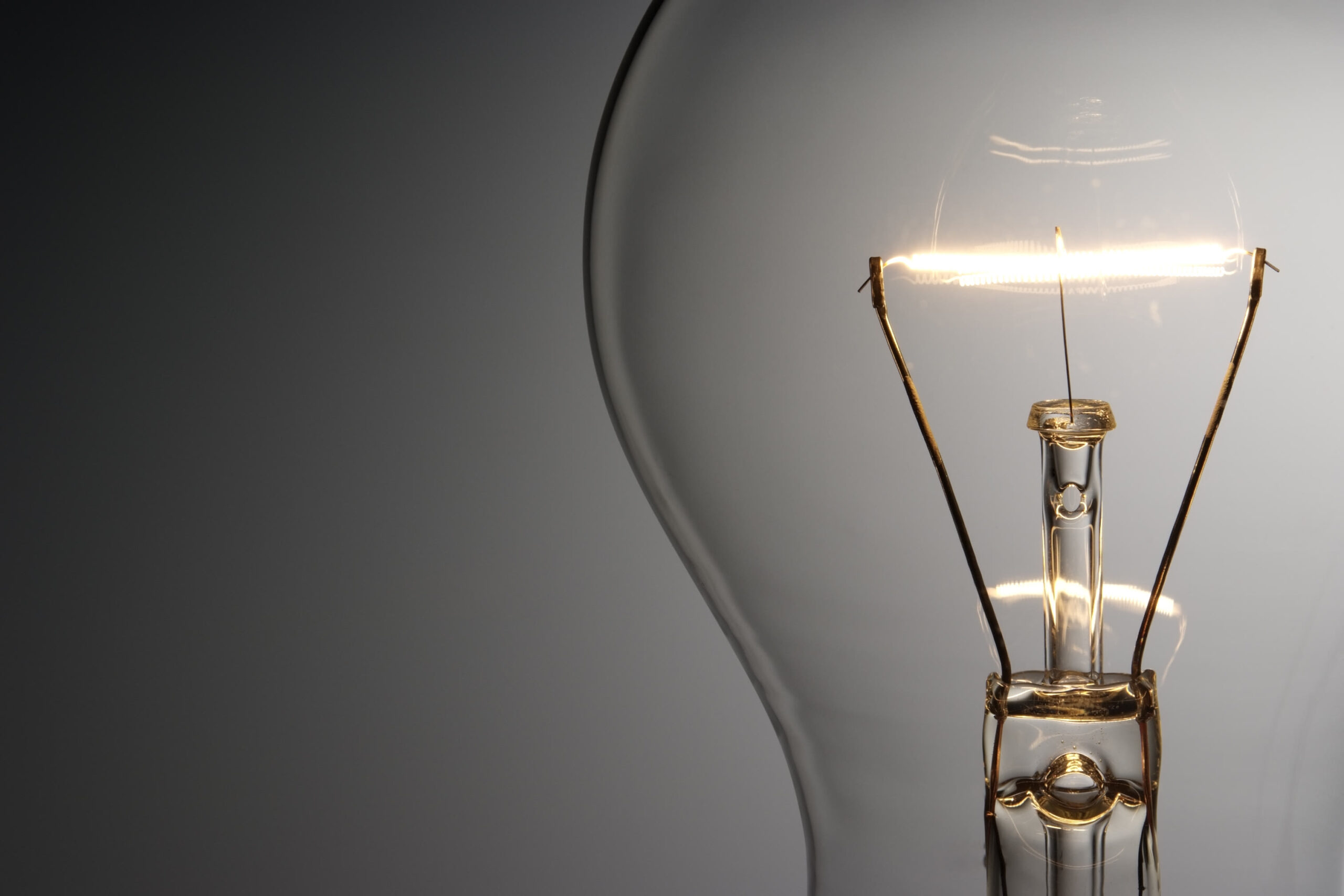 Close-up of a clear light bulb with a visible glowing filament, set against a plain gray background. The left side of the bulb is out of frame, and the image has a soft gradient lighting.