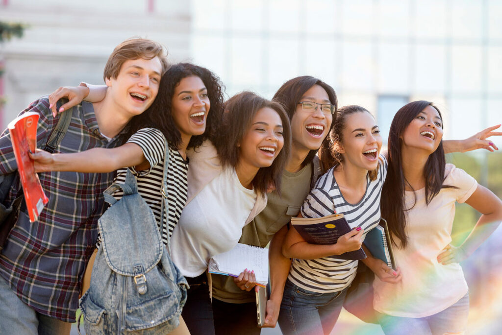 A group of six smiling young adults stand close together outdoors, some holding books and backpacks, looking and pointing excitedly toward something in the distance.