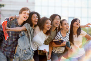 A group of six smiling young adults stand close together outdoors, some holding books and backpacks, looking and pointing excitedly toward something in the distance.