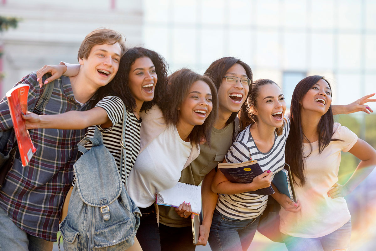 A group of six smiling young adults stand close together outdoors, some holding books and backpacks, looking and pointing excitedly toward something in the distance.