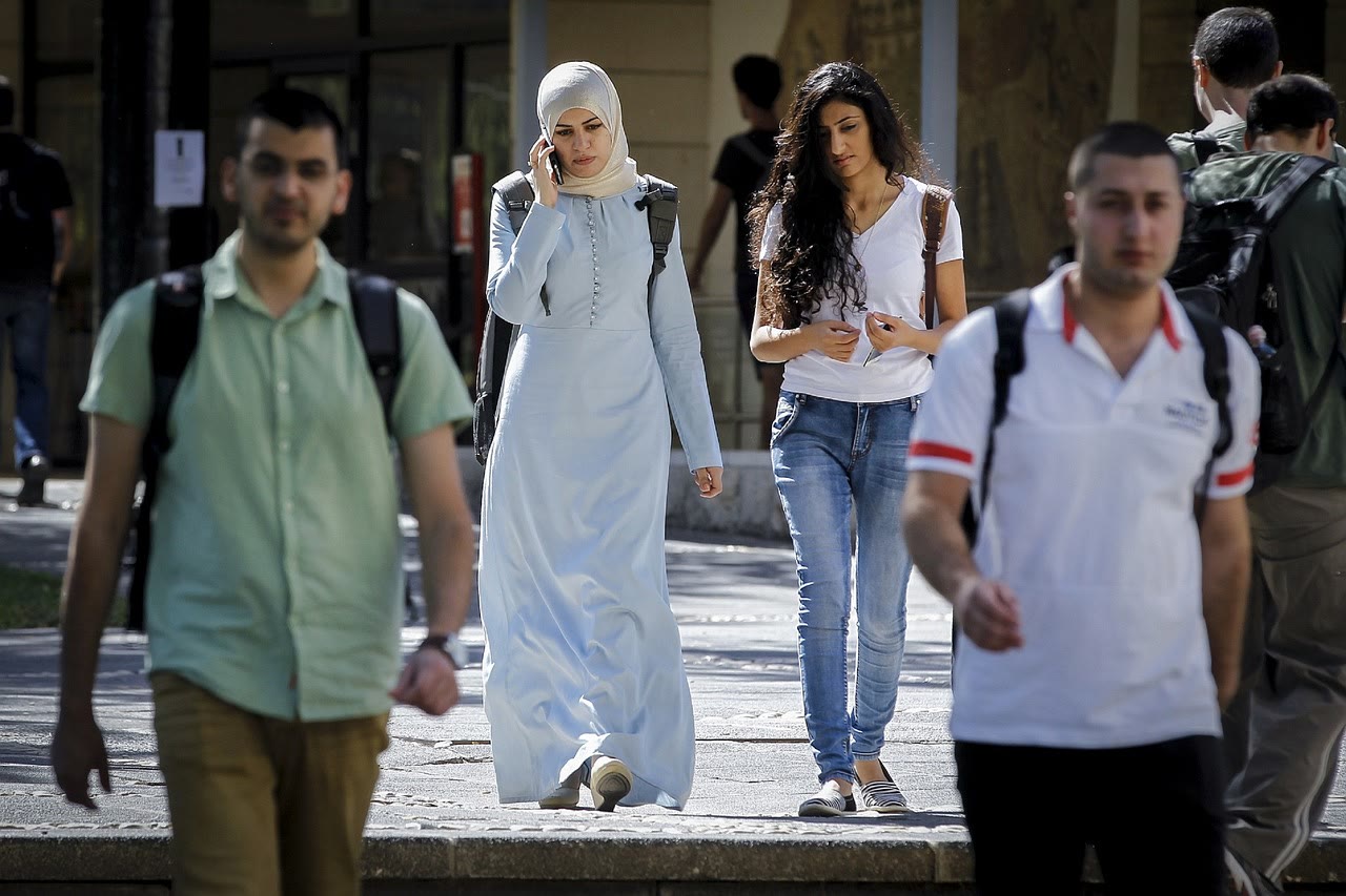 Several young adults walk outdoors. A woman in a light blue dress and hijab talks on her phone, while another woman in a white shirt and jeans walks beside her. Other people with backpacks walk in different directions.