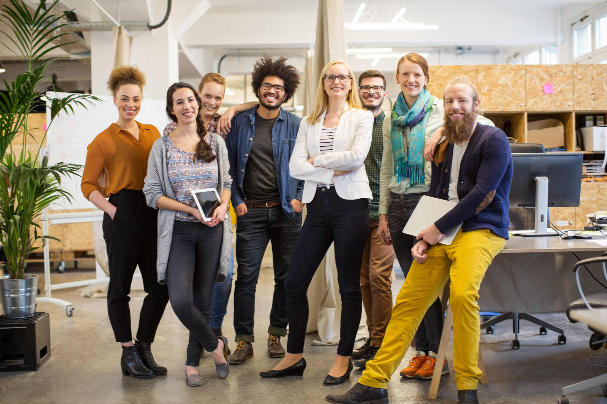 A group of eight people, casually dressed, stand and smile together in a modern, open-plan office space with computers, plants, and a relaxed atmosphere.