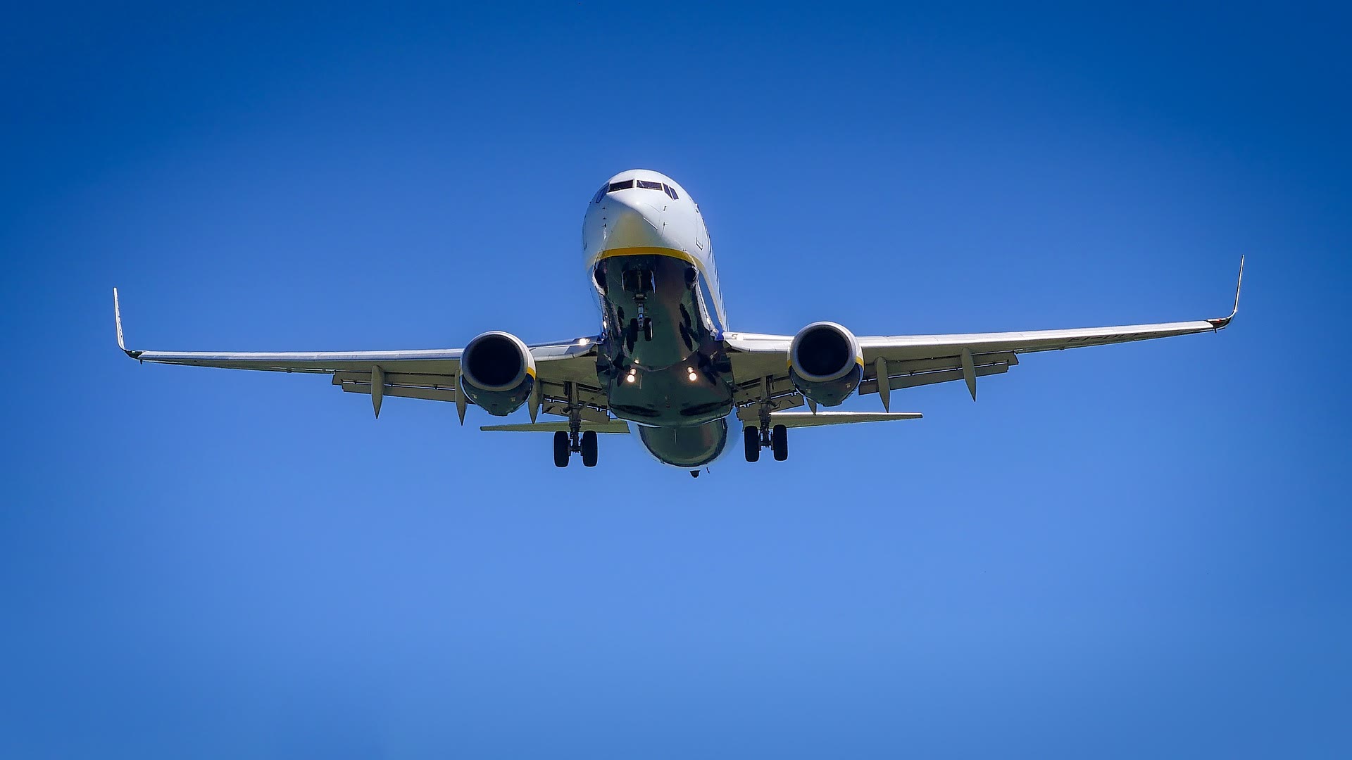 A commercial airplane viewed from below, flying against a clear blue sky with its landing gear extended and both engines visible.