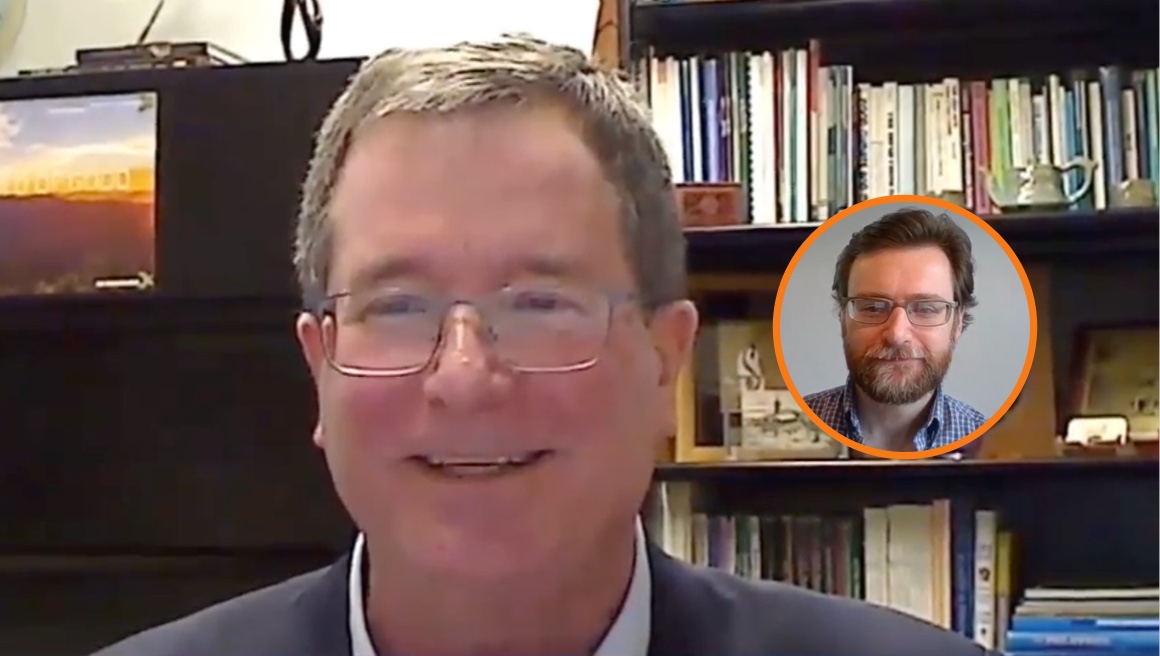 A man with glasses smiles in front of a bookshelf during a video call. In the corner, there is a circular inset of another man with glasses and a beard, also smiling, against a blurred background.