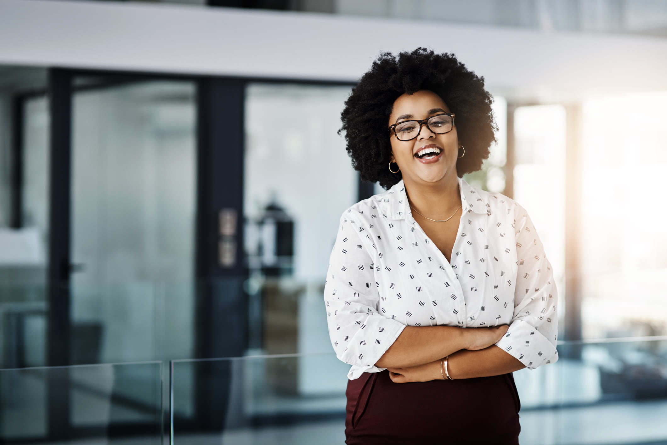 A woman with curly hair and glasses, wearing a white patterned shirt and dark pants, stands indoors smiling with her arms crossed. The background shows modern office decor and large glass windows.