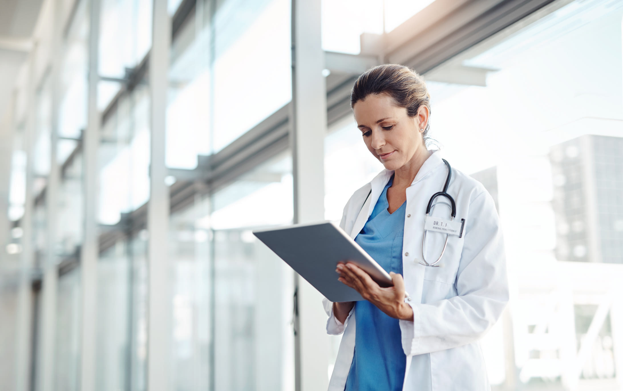 A doctor wearing a white coat and stethoscope stands in a bright, modern hospital hallway, looking at a digital tablet.